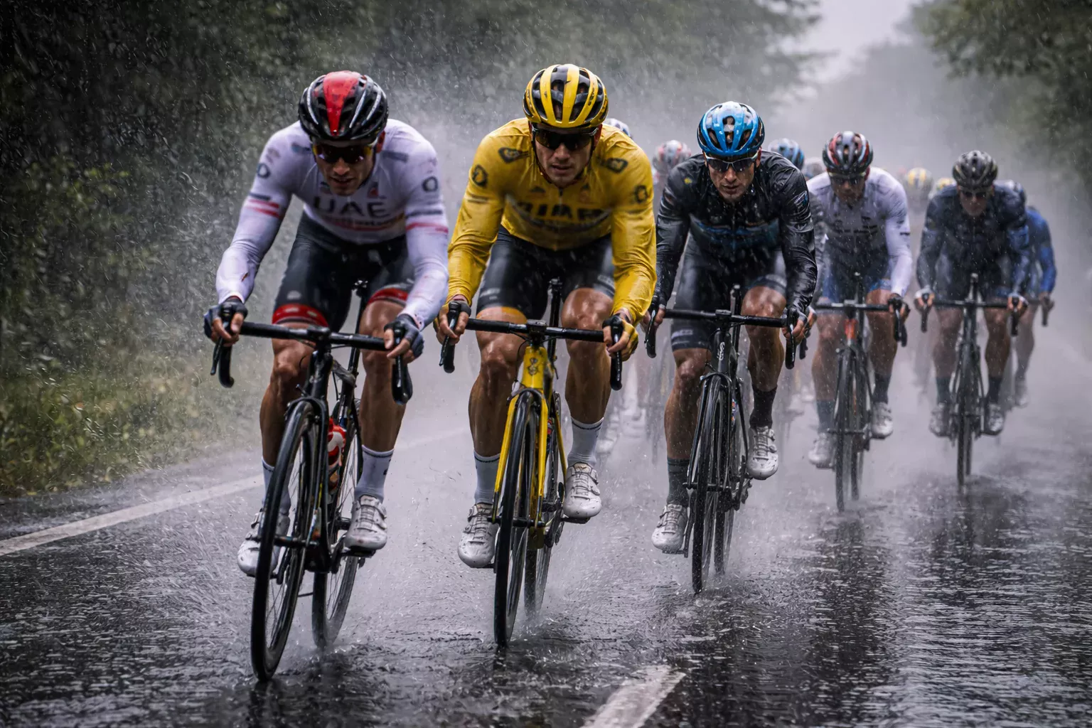 Pelotón de ciclistas rodando bajo lluvia intensa con la carretera mojada y spray de agua